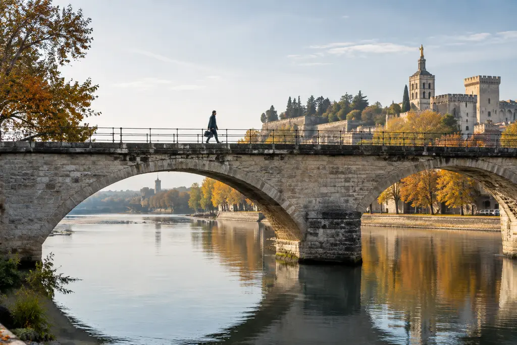 Un passant traverse un pont de pierre au-dessus d’une eau calme à Avignon.
